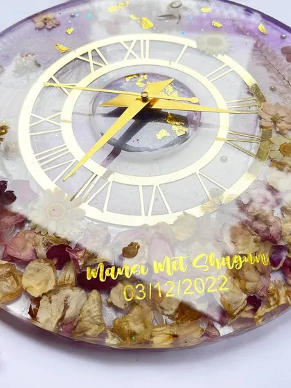 This angle highlights the glossy finish and the way the light reflects off the gold details and the preserved flowers in this lavender clock.