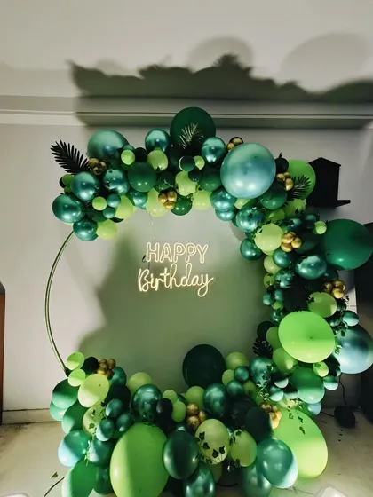 A lush, monochrome green balloon arch on a ring frame, accented with gold chrome balloons and a 'Happy Birthday' neon sign.