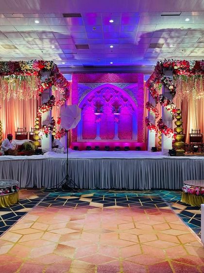 A grand stage setup inside a banquet hall, featuring two mandap-style structures on either side of a central backdrop with purple and pink lighting.
