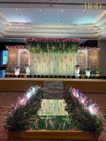 A grand stage with a lush green and floral canopy, a backdrop of hanging flowers, and a unique water feature at the front of the aisle.