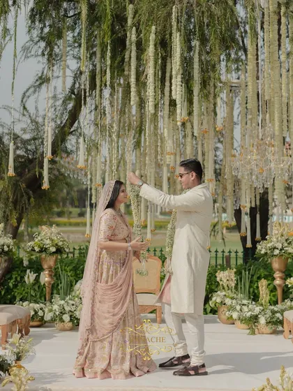 The Jaimala ceremony begins. The groom playfully bows as the bride places the garland, a moment of joy captured against the beautiful backdrop of hanging florals.