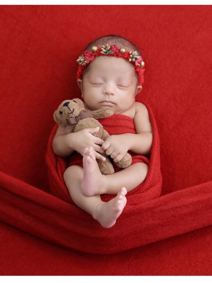 A sweet portrait of a sleeping baby girl in a red wrap, cuddling a tiny teddy bear. The simple setup allows her peaceful expression to be the main focus.