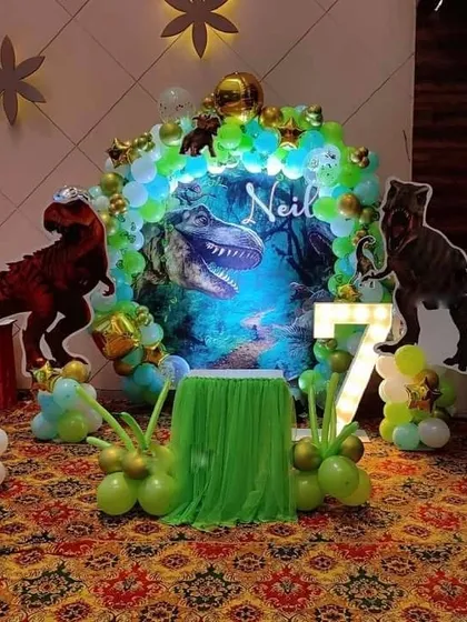 A thrilling dinosaur-themed 7th birthday party. The setup includes a jungle backdrop, large T-Rex cutouts, and a '7' marquee light.