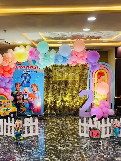 A complete Cocomelon birthday setup in a party hall. This decoration features a large backdrop with all the Cocomelon characters, a gold sequin panel, character cutouts, and a pastel rainbow balloon arch.