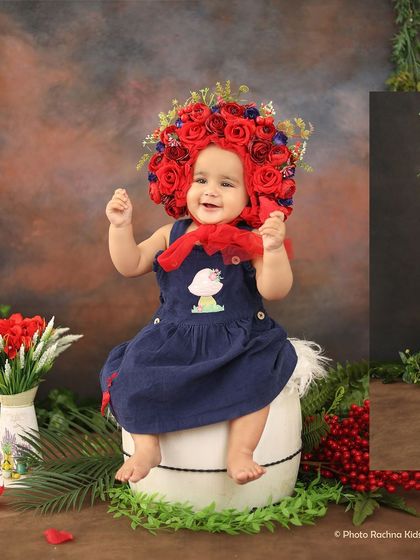 A happy baby girl wearing a stunning red rose bonnet, sitting in a setup with lush greenery.