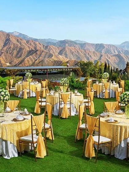 A stunning outdoor reception setup with a mountain backdrop. The gold-themed tables and chairs look incredibly luxurious against the green lawn.