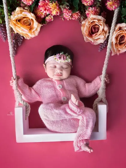 This sweet newborn girl is dressed in a pink knitted outfit, sleeping on a miniature white swing adorned with flowers. The vibrant pink background makes this a striking and memorable shot.