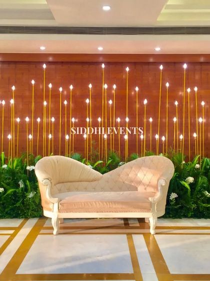 This modern stage design uses a backdrop of glowing vertical rods and a bed of lush greenery to create a unique and sophisticated setting for the couple's chaise lounge.
