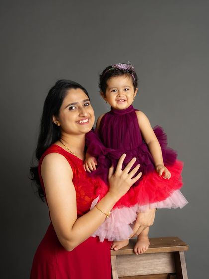 A mother holds her smiling daughter in a sweet studio portrait. The coordinating red and purple outfits add a pop of color and fun to the image.