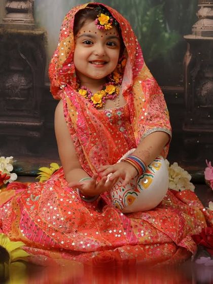 With a pot of makhan and a sweet smile, this little Radha is the picture of grace. The vibrant colors of her outfit and the floral setting create a stunning and traditional portrait.
