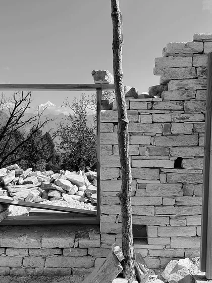 A black and white photograph capturing the raw beauty of the construction process. A window frame perfectly captures the distant mountains, turning a pile of stones into a piece of emergent architecture.