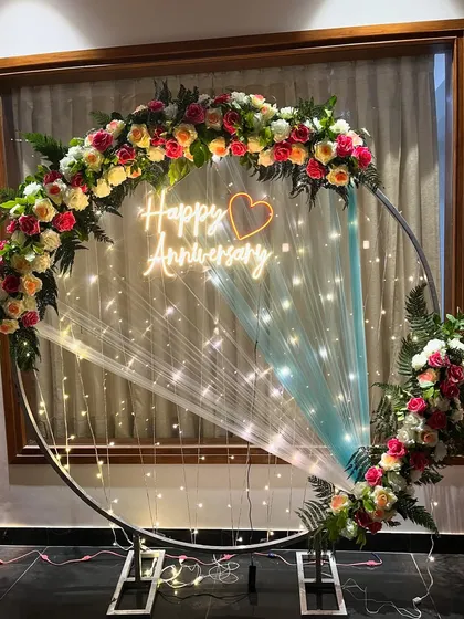 An elegant ring backdrop perfect for an anniversary. This setup features a metal ring decorated with fresh, colorful flowers, delicate fairy lights, and a custom 'Happy Anniversary' neon sign, creating a beautiful and romantic focal point.