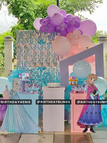 This Frozen party setup features a blue and purple shimmer wall, creating a sparkling, icy effect. Life-sized cutouts of Anna and Elsa stand beside themed plinths, perfect for a royal celebration.