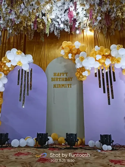 This elegant stage setup features lavender arched panels with gold and white balloon decor. The overhead floral arrangement and stage lights create a grand and luxurious feel for a birthday celebration.