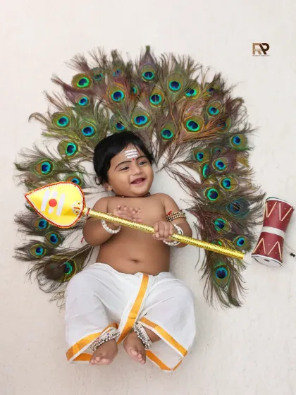 A creative and divine portrait of a baby dressed as Lord Murugan, lying on a bed of peacock feathers and holding a vel. The joyful expression makes this a truly special image.