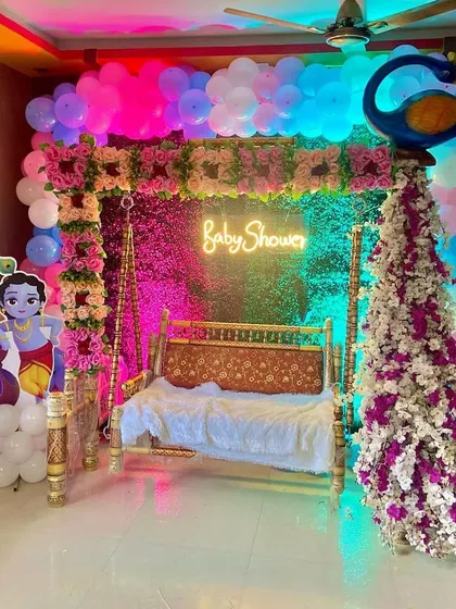 An indoor baby shower setup with a Radha-Krishna theme, featuring a decorated swing, character cutouts, and a colorful balloon arch.