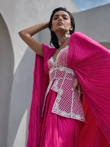 A vivid, hot pink ensemble featuring an embellished corset top paired with a draped skirt and a dramatic, crushed fabric cape. The look is a play on structure and fluidity.
