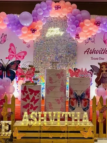 A butterfly and fairy theme with a wooden picket fence and crate props. The purple and peach balloon arch creates a warm and inviting atmosphere.
