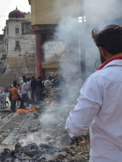 A man stands watching the pyres, his back to the camera. This shot focuses on the personal, contemplative side of being at the ghats, a moment of reflection amidst the smoke.
