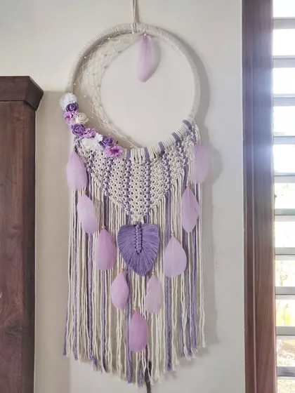Spread like wildflowers. This crescent moon macrame dreamcatcher in shades of lavender and off-white is a delicate and whimsical piece, adorned with feathers and a single macrame leaf.