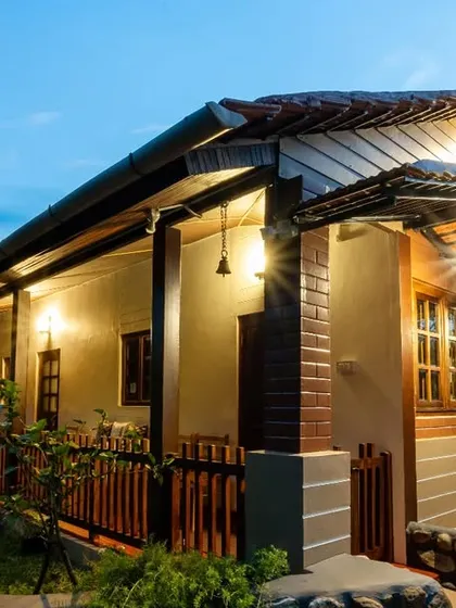 The exterior of the "Eorthe" homestay in Coorg at twilight. The warm lights from the cottage create a welcoming glow, making it look like a cozy and peaceful retreat.
