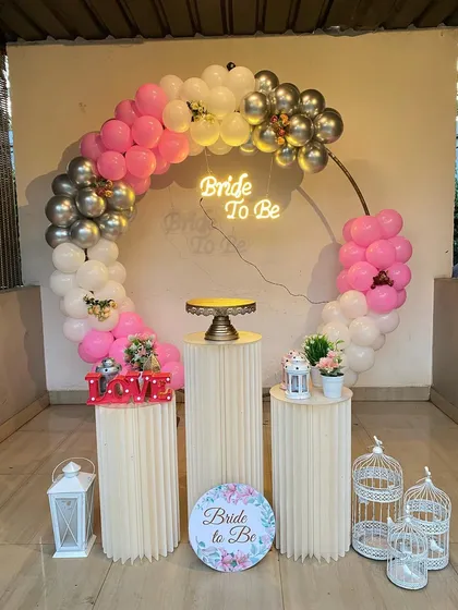 Here is a fun and vibrant bachelorette party setup. The centerpiece is a circular frame with a multi-colored balloon arch in pink, white, and silver chrome. A 'Bride to Be' neon sign adds a fun glow, while decorative plinths, lanterns, and birdcages complete the theme.