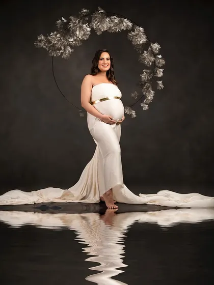 An elegant portrait against a dark background, framed by a silver floral hoop. The simple white gown and water reflection create a sophisticated and artistic composition.