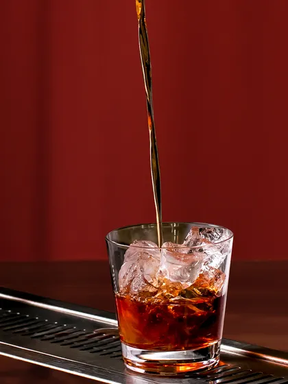 The perfect pour. A stream of rich, dark liquor cascades over ice, a simple yet satisfying moment in the creation of a classic cocktail on the rocks.