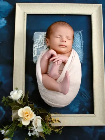 A newborn baby is swaddled in white and posed inside a picture frame against a floral blue backdrop, creating a beautiful "living art" portrait.