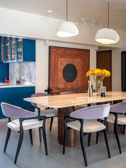 The dining area is designed for memorable gatherings, featuring a striking marble-topped table with sculptural wooden legs. We used lavender-hued chairs to introduce a soft, sophisticated color that complements the bold blue of the adjacent open kitchen.