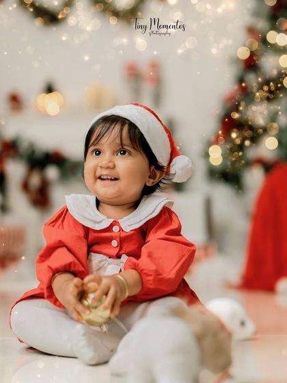 The littlest Santa. Her happy expression and festive hat, set against a backdrop of twinkling lights, make for a perfect holiday photo.
