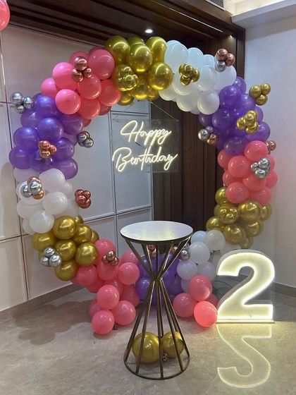 A vibrant balloon ring decoration for a second birthday. The multi-colored garland of pink, purple, gold, and white balloons is paired with a 'Happy Birthday' neon sign and a large light-up number '2'.