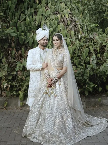 A classic wedding portrait of the couple posing against a lush green backdrop, showcasing the intricate details of their white wedding outfits.