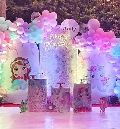 An outdoor mermaid theme birthday party for Andy, set at dusk. The colorful balloon arch and character cutouts are illuminated, creating a magical underwater glow for the evening celebration.