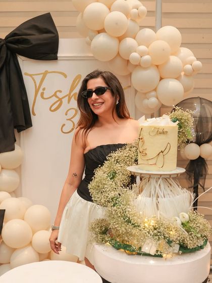 The birthday girl posing with her stunning cake display. We designed this chic setup with a baby's breath floral cascade, tying into the elegant black and white bow theme of her 35th birthday party.