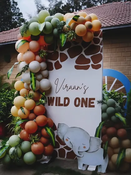 A "Wild One" welcome sign for Viraan's first birthday. The sign is framed by a lush balloon garland in earthy tones of green, orange, and brown, with a cute elephant prop.