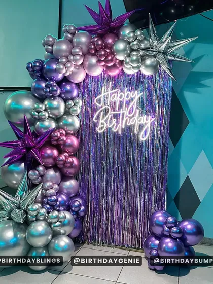 A vibrant setup with a purple fringe backdrop and a neon "Happy Birthday" sign. The balloon arch in shades of purple and silver, with starburst foil balloons, creates a futuristic, high-energy vibe.