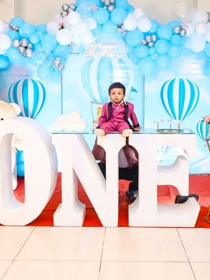 A grand first birthday celebration with a hot air balloon theme. The setup features large 'ONE' letters, a beautiful blue and white balloon arch, and adorable teddy bear accents, creating a picture-perfect scene for the birthday boy.