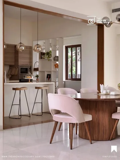 The dining area in 'A Walk in the Clouds', with a fluted pedestal table and blush pink chairs, is visually connected to the kitchen by a large, smoked glass partition.