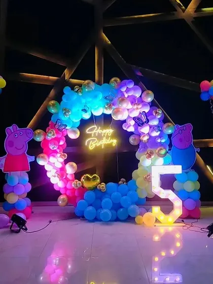A colorful Peppa Pig theme decoration for a 5th birthday, with a rainbow balloon arch, marquee number '5', and character cutouts.