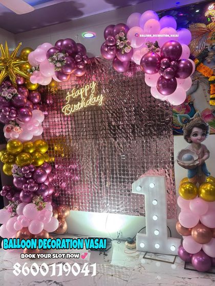 A beautiful first birthday setup with a Little Krishna theme. The decoration includes a pink and chrome-gold balloon arch against a shimmer backdrop, a neon sign, and a character cutout.