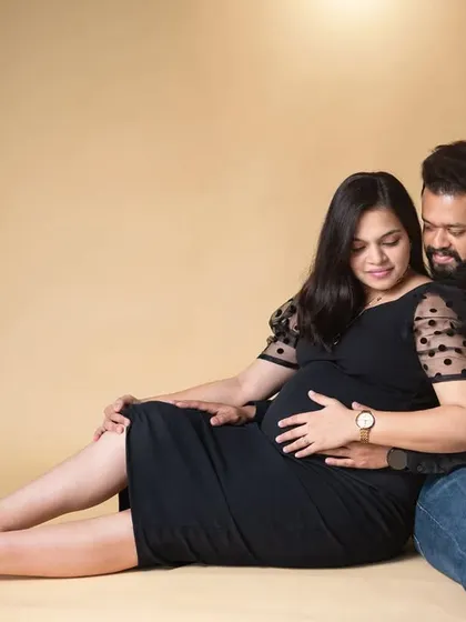 A relaxed, full-length portrait of an expecting couple seated on the floor against a neutral beige backdrop. This image captures a comfortable and natural moment of togetherness during their studio maternity session.