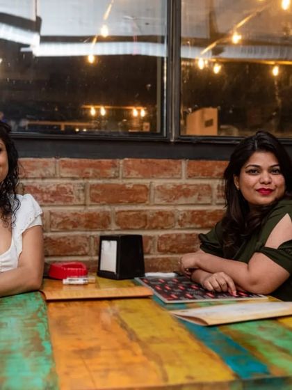 Two friends enjoying a quiet moment at one of our colorful tables. The rustic brick wall provides a warm and inviting backdrop.
