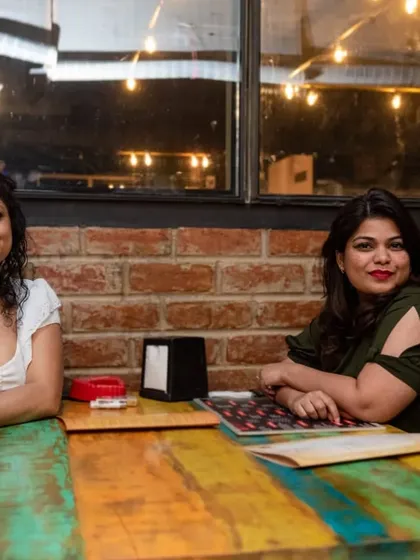 Two friends enjoying a quiet moment at one of our colorful tables. The rustic brick wall provides a warm and inviting backdrop.