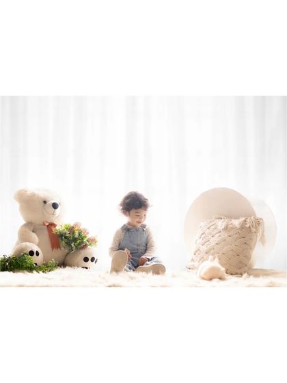 A toddler sits quietly on a fluffy rug, surrounded by a large teddy bear and other soft props. This calm and gentle studio scene captures a moment of peaceful play.