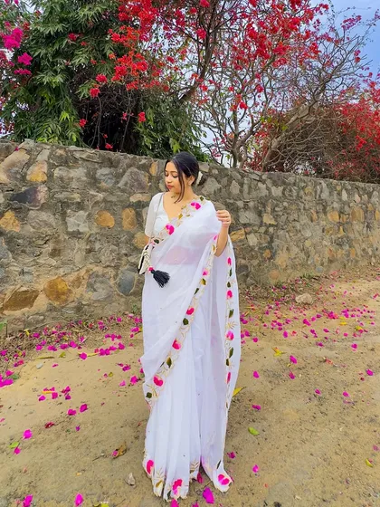 A full-length view of the white mul cotton saree against a stone wall and bougainvillea. The floral embroidery on the saree border complements the natural surroundings perfectly.