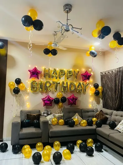 A simple yet festive birthday decoration for a living room. The setup features 'Happy Birthday' foil balloons against the wall, with yellow and black balloons scattered around.