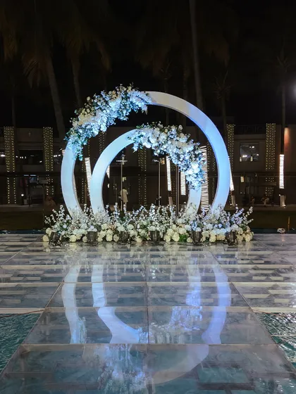 A modern and elegant stage setup for a reception or sangeet. The interlocking white rings, adorned with white florals, are placed on a transparent stage built over a pool, creating a stunning reflection and a floating effect.