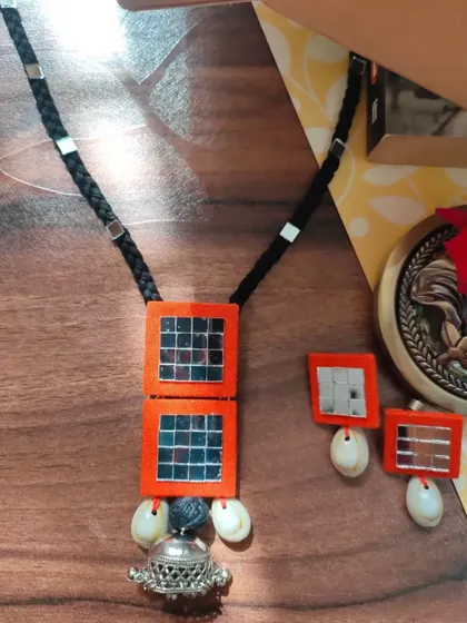 A complete view of this handmade Navratri necklace set. It features bright orange fabric squares with intricate mirror work, cowrie shell accents, and a large oxidized silver jhumka pendant.