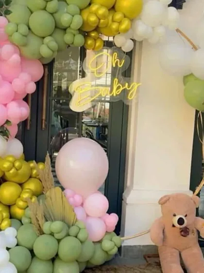 A close-up look at an 'Oh Baby' entrance decoration. This arrangement features a mix of pastel green, pink, and yellow balloons, a neon sign, and a welcoming teddy bear prop.